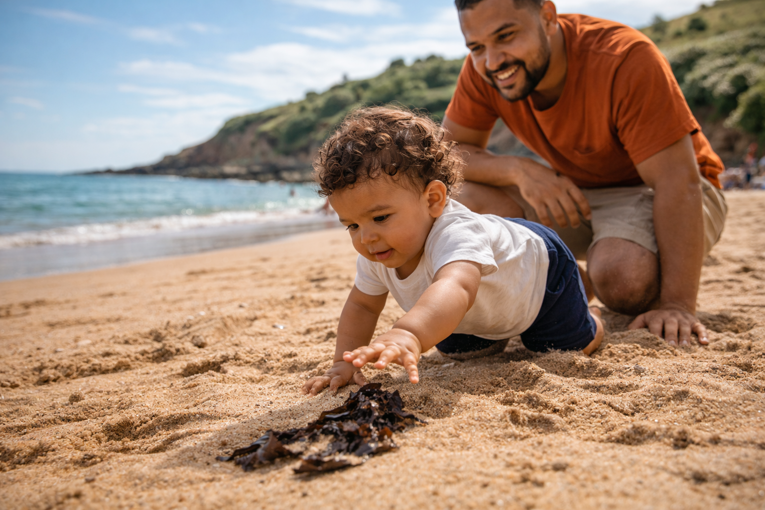 A 9-month-old baby crawling on a sandy UK beach, reaching for a shell, parent crouching nearby watching closely on a bright summer day