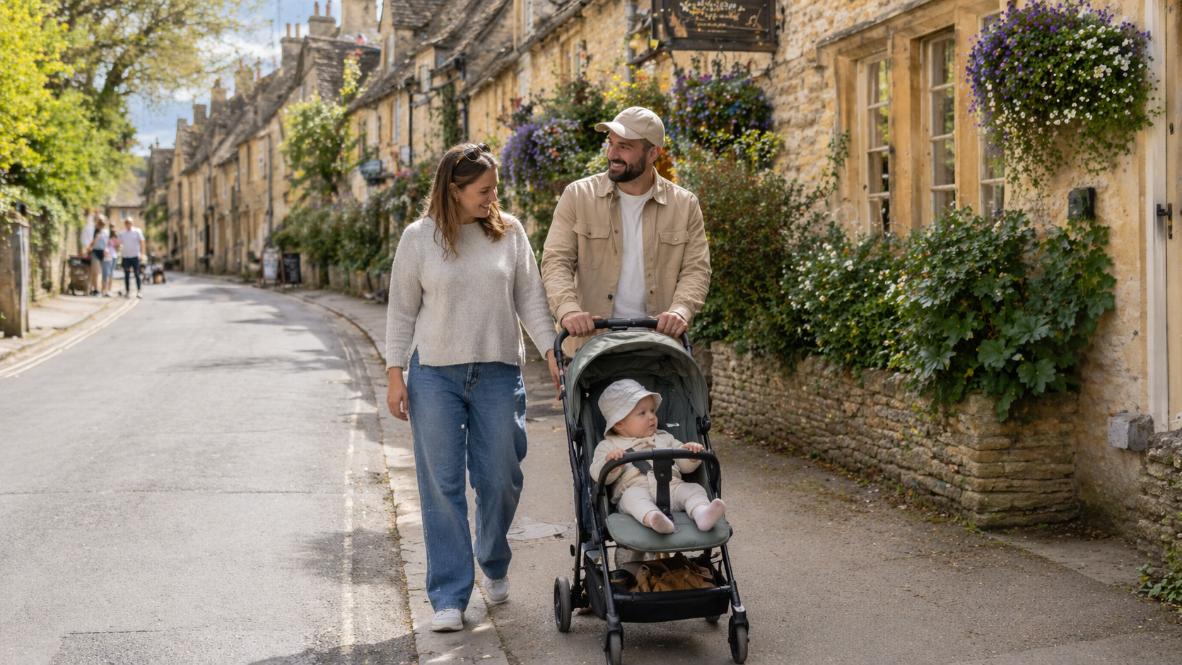 Parents pushing a baby in a pushchair along a honey-stone Cotswolds village street on a bank holiday weekend break