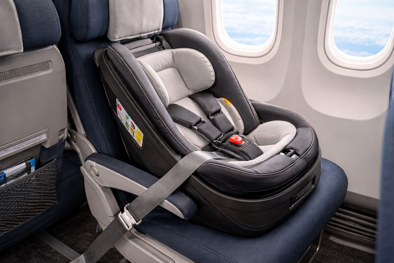 A rear-facing infant car seat properly installed on an aircraft passenger seat, aircraft seatbelt securing the car seat base, cabin interior visible