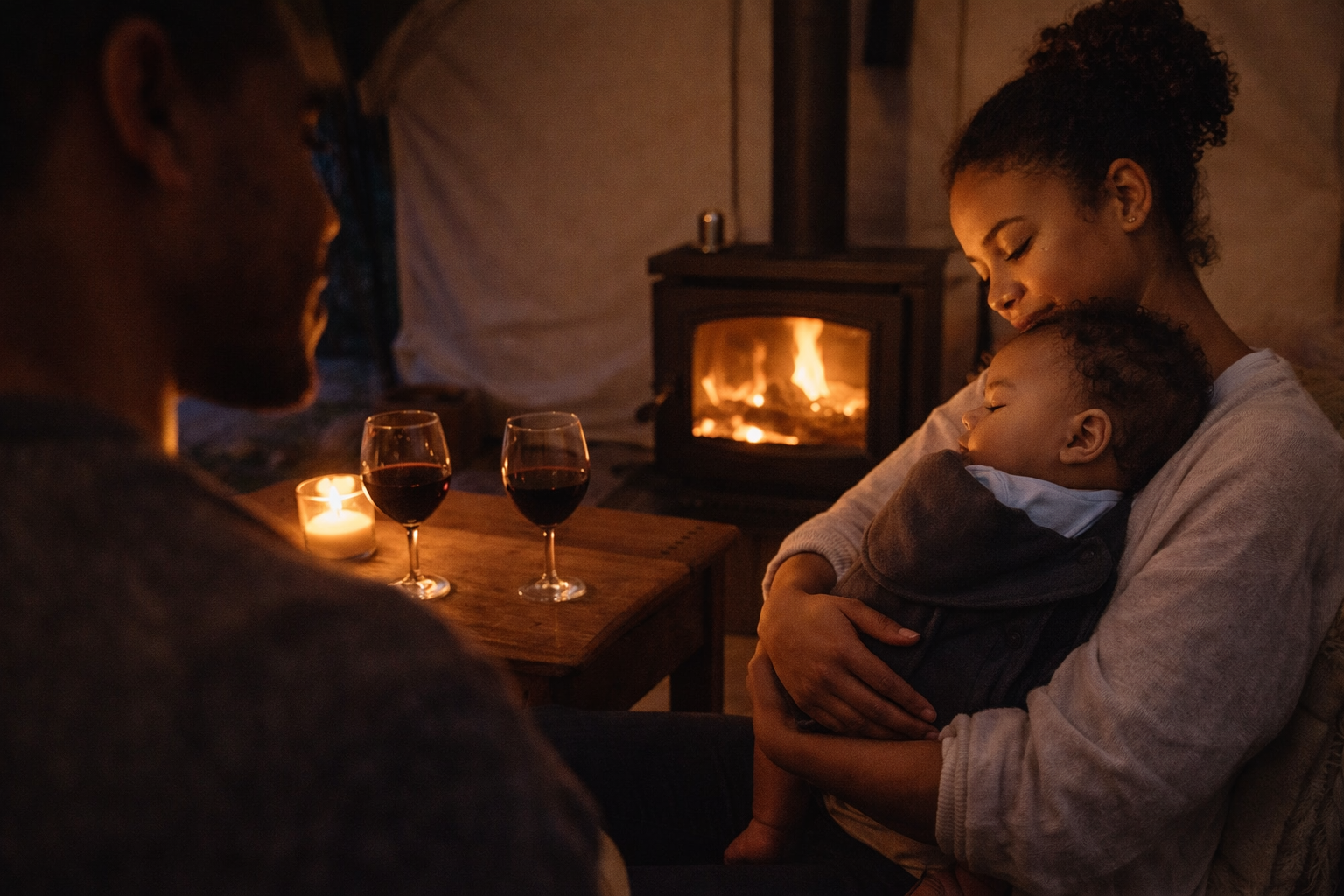 A family in a cosy cottage in the evening — fire glowing, baby asleep on a parent's chest, wine on the table, the relaxation that actually works with a baby